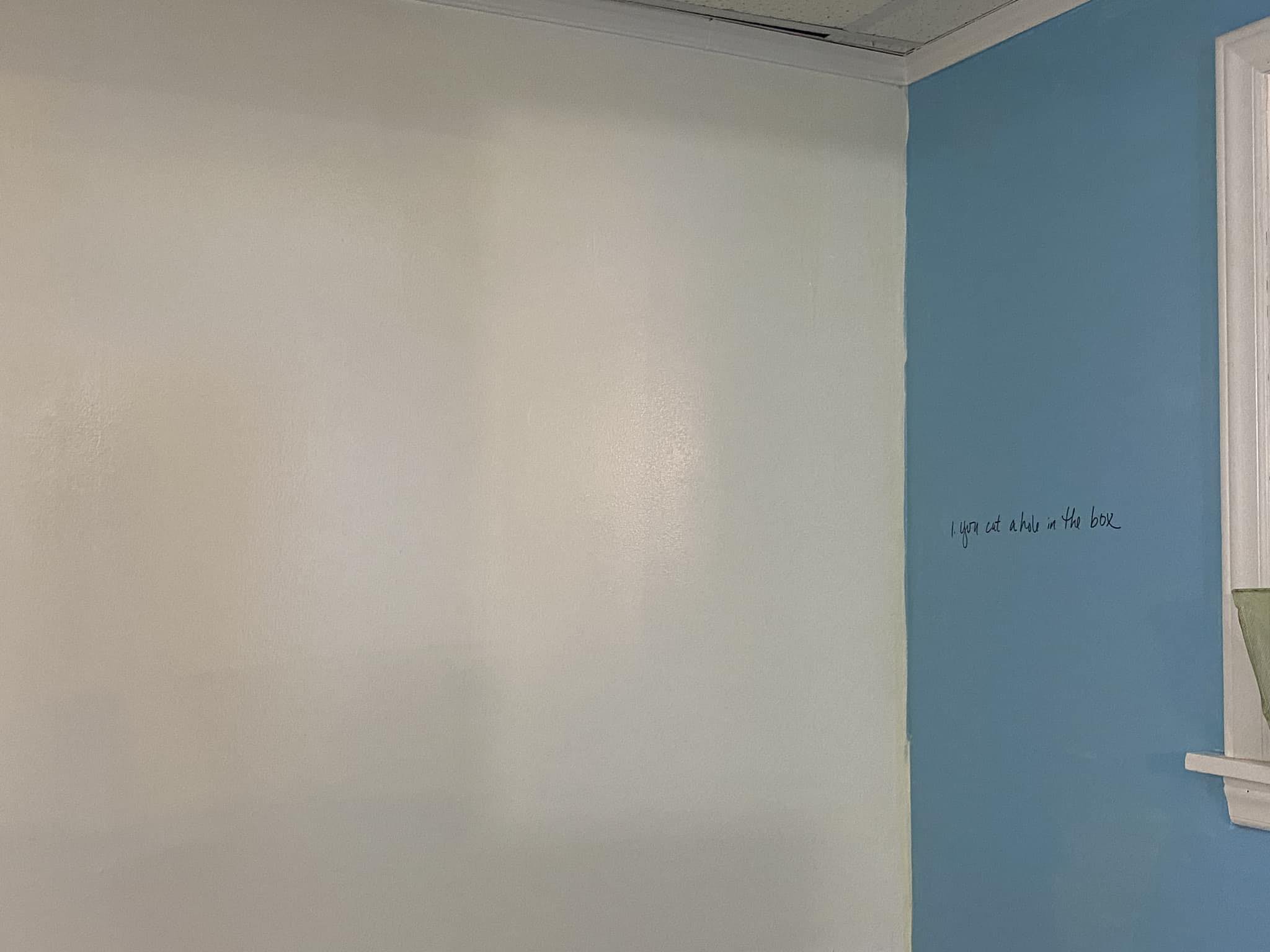 A corner wall, one side white, the other blue with a dry erase sentence that reads: 1. You cut a hole in the box.